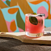A pink cocktail with a black olive sits on a wooden board, lime wedges and crumbs beside it, against a colorful mural backdrop.
