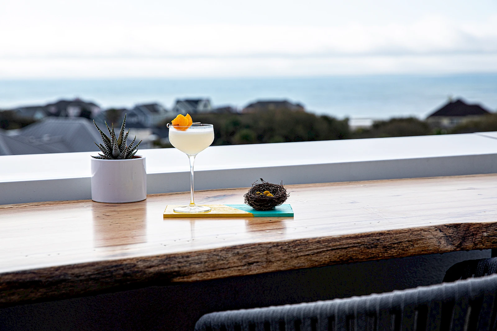 A seaside bar scene with a cocktail, a small dessert on a napkin, and a potted plant on a wooden counter overlooking the ocean, classy vibes.