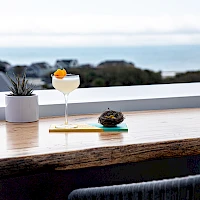 A seaside bar scene with a cocktail, a small dessert on a napkin, and a potted plant on a wooden counter overlooking the ocean, classy vibes.