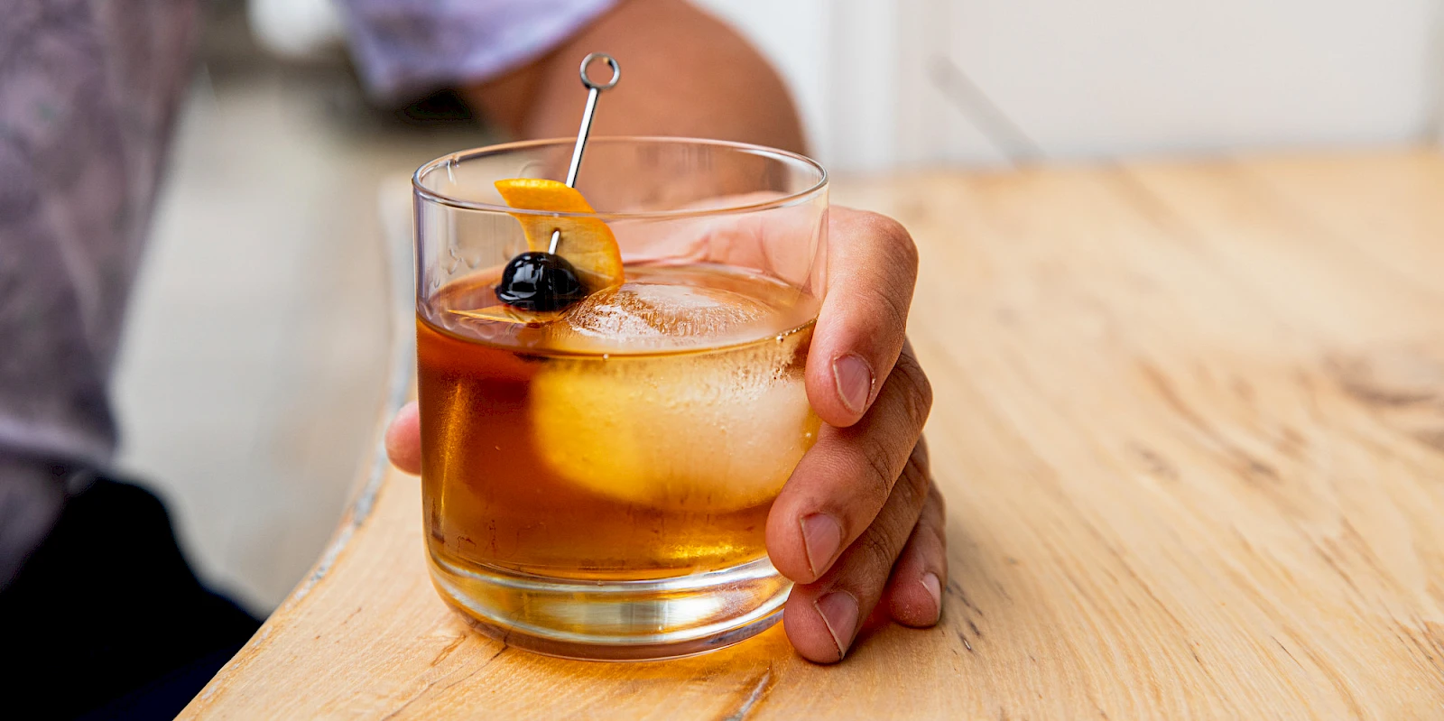 A whiskey or bourbon on the rocks in a short glass, with an orange twist and a dark cherry garnish on a metal pick, resting on a wooden table.