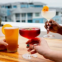 A sunny rooftop cheers: four colorful cocktails clink in a toast as hands hover over a wooden table with a backdrop of city buildings.