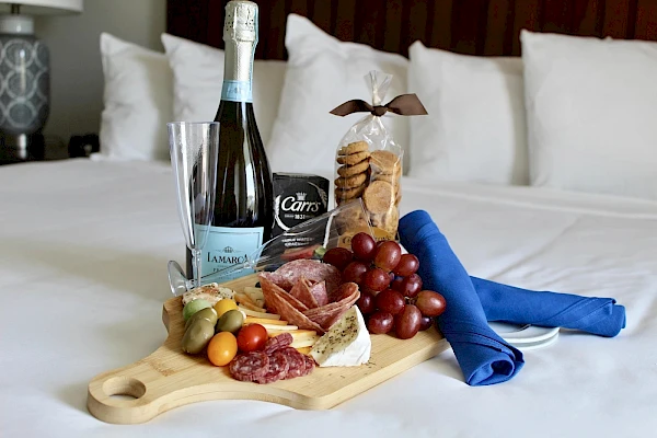 A breakfast-in-bed setup with a wooden board of cheeses, grapes, and crackers, plus a small pastry, a bottle of wine, a glass, and a blue napkin on a white bed.