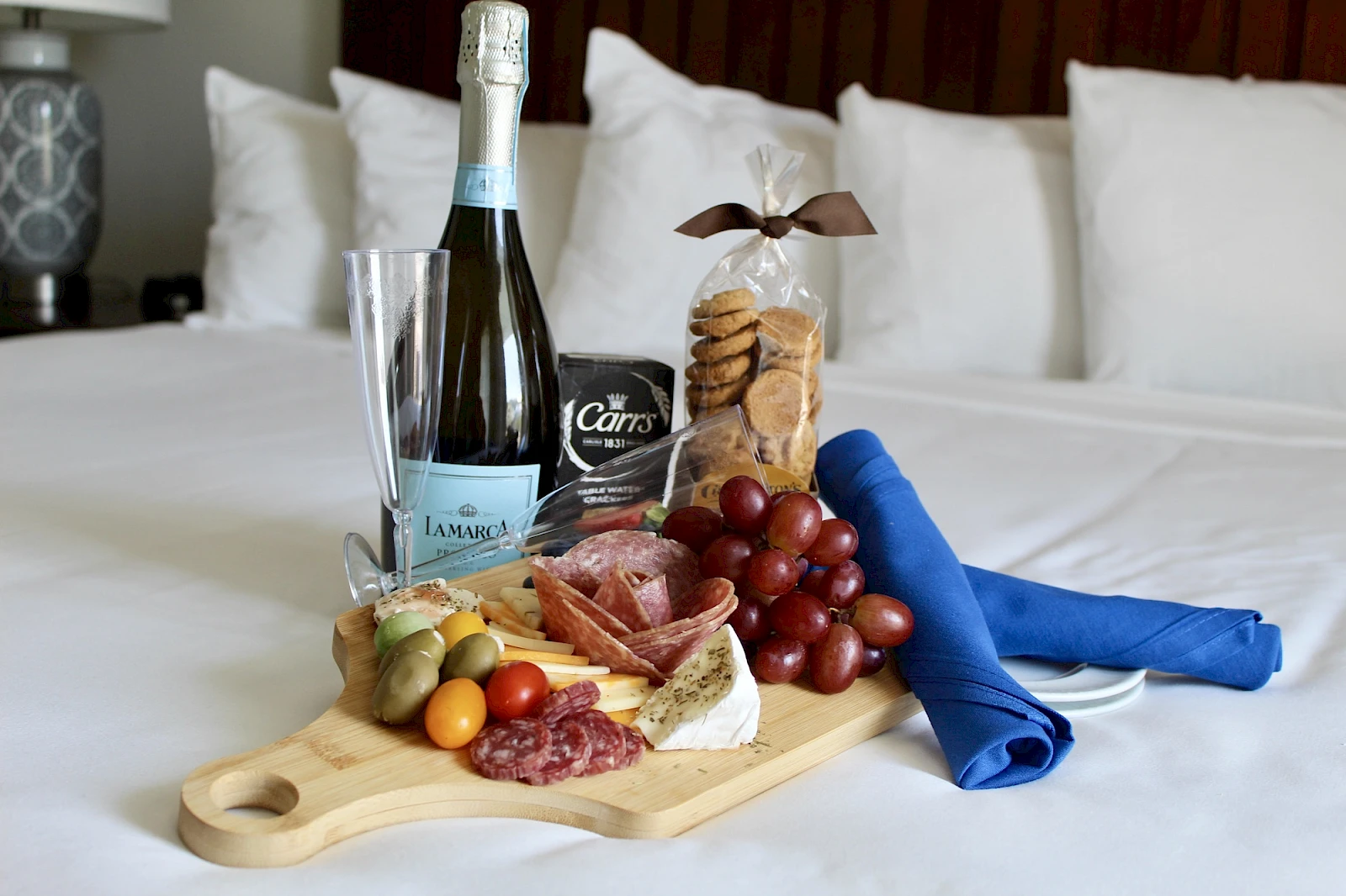 A wine bottle, a glass, crackers, cheese, grapes, fruits on a wooden board with a blue napkin and a snack bag on a bed.
