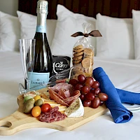 A wine bottle, a glass, crackers, cheese, grapes, fruits on a wooden board with a blue napkin and a snack bag on a bed.