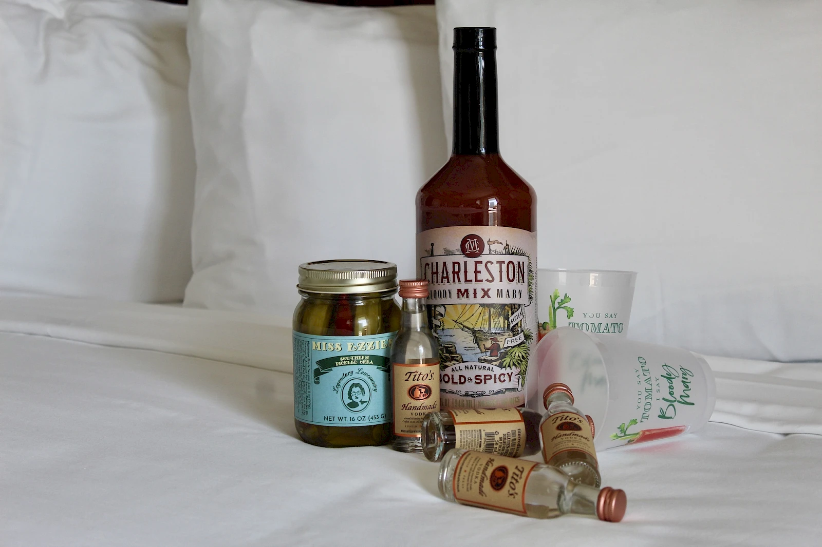 Assorted condiments on a bed: a whiskey bottle, jars of pickles, hot sauce, small spice jars, and a few spice sachets, all resting on white sheets.