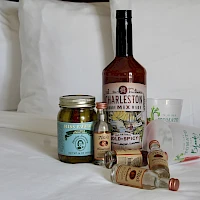 Assorted condiments on a bed: a whiskey bottle, jars of pickles, hot sauce, small spice jars, and a few spice sachets, all resting on white sheets.