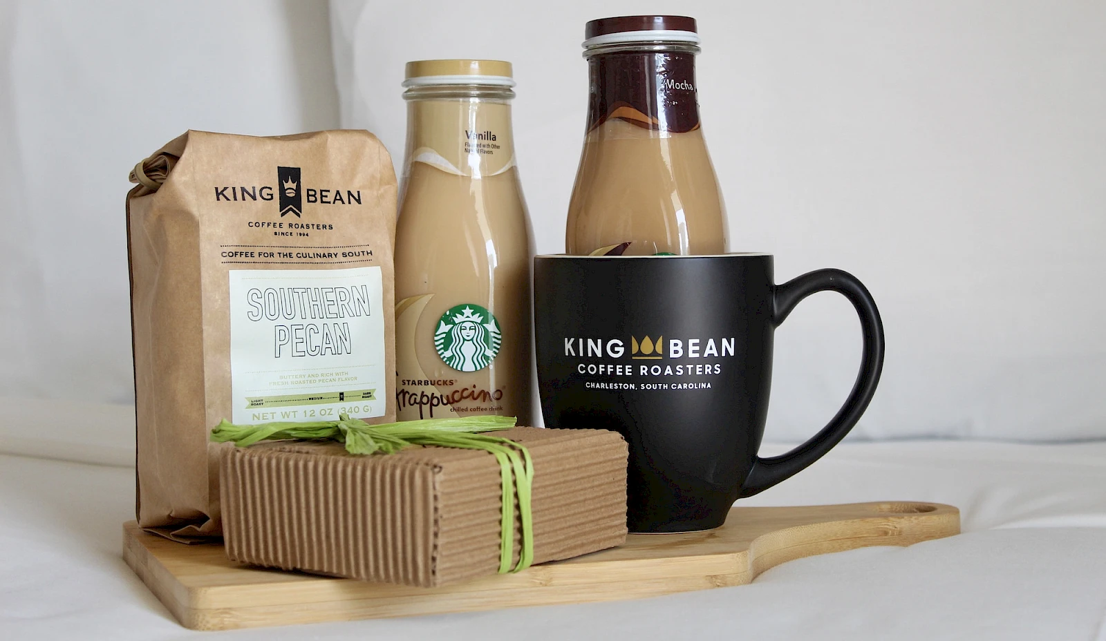 A coffee gift set: bag of beans, two bottles (milk/creamer), a mug reading “King Bean Coffee Roasters,” and a wrapped treat on a wooden tray.