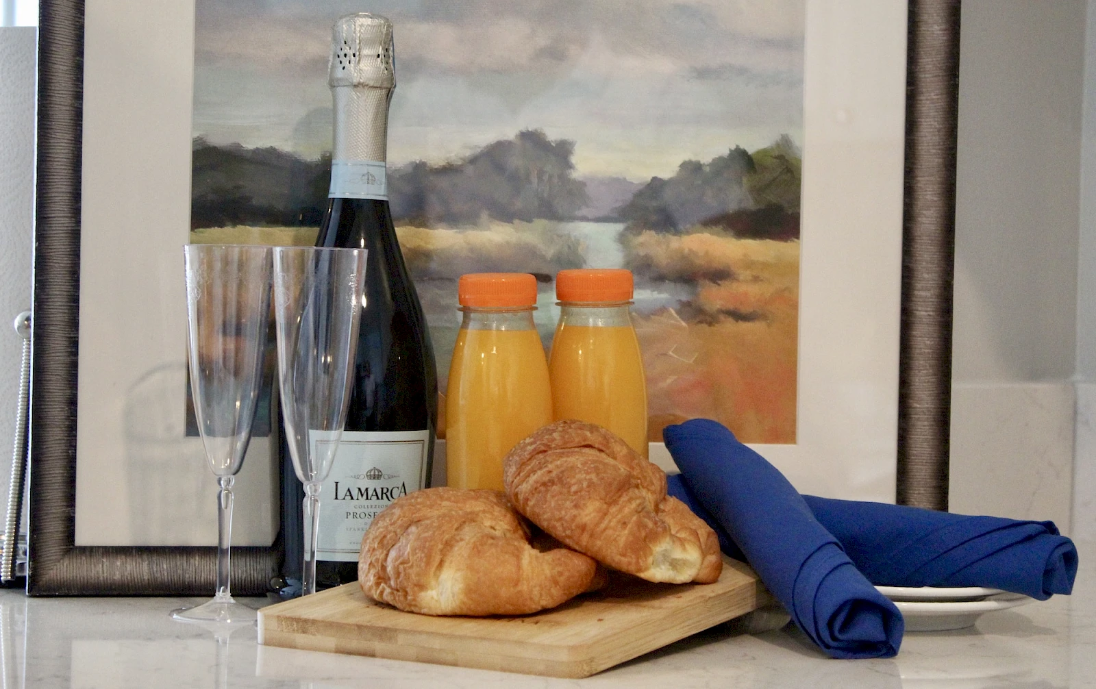 A classy breakfast setup with sparkling wine, orange juice, two croissants, and blue napkins on a wooden board.
