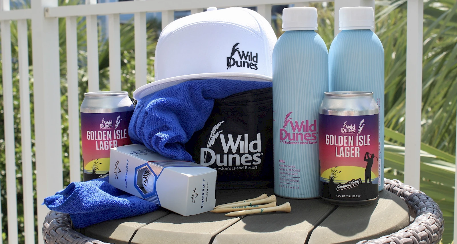 A beachy outdoor cooler setup with Wild Dunes gear, blue towels, two light blue water bottles, cans of Golden Isle Lager, sunglasses case, and a pineapple-pattern box.