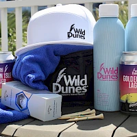 A beachy outdoor cooler setup with Wild Dunes gear, blue towels, two light blue water bottles, cans of Golden Isle Lager, sunglasses case, and a pineapple-pattern box.