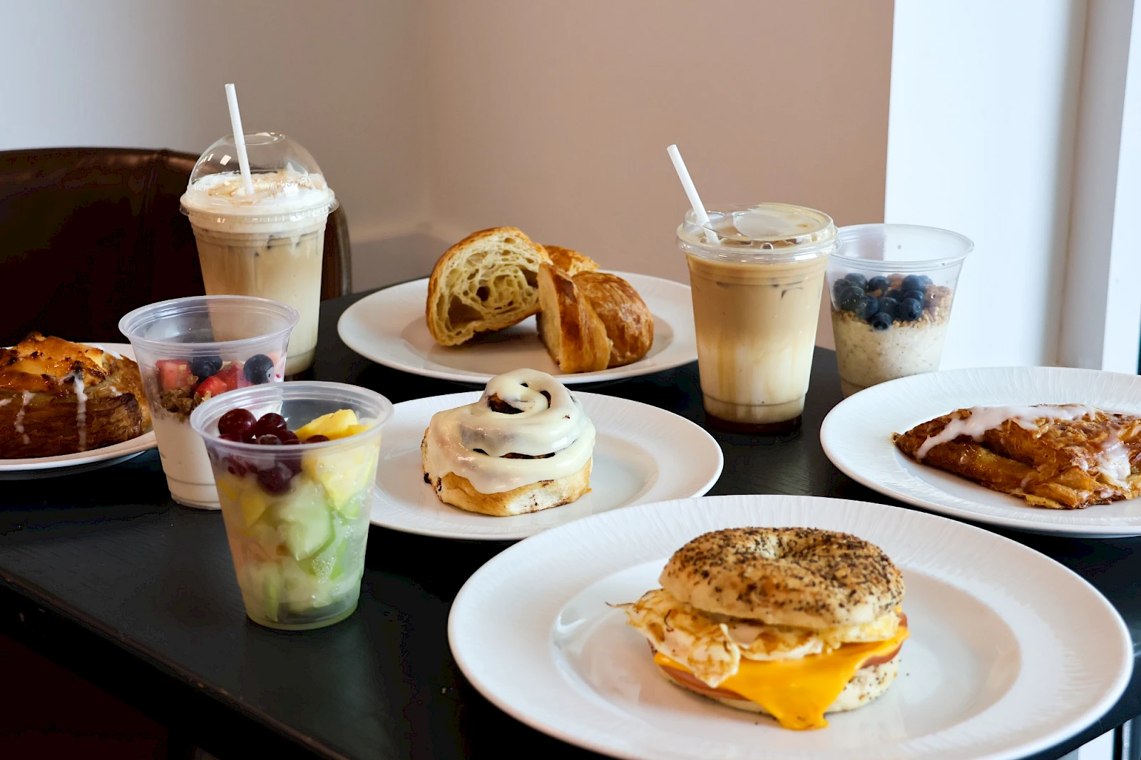 A spread of breakfast treats: pancakes, muffins, bagels, croissants, yogurt cups, fruit cups, oatmeal cookies, and coffee drinks on a table.