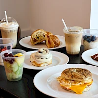 A spread of breakfast treats: pancakes, muffins, bagels, croissants, yogurt cups, fruit cups, oatmeal cookies, and coffee drinks on a table.