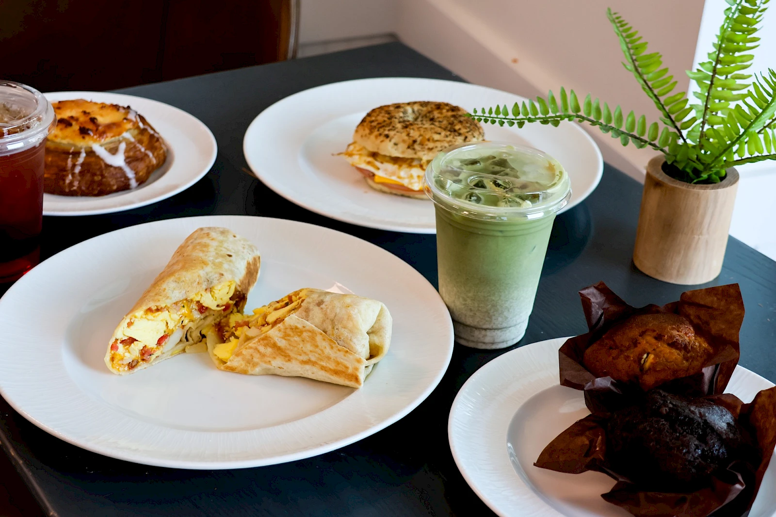 A spread of breakfast items: ham and cheese pastry, veggie wrap, green tea latte, iced matcha, and chocolate brownies on plates with a fern plant nearby.
