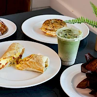 A spread of breakfast items: ham and cheese pastry, veggie wrap, green tea latte, iced matcha, and chocolate brownies on plates with a fern plant nearby.