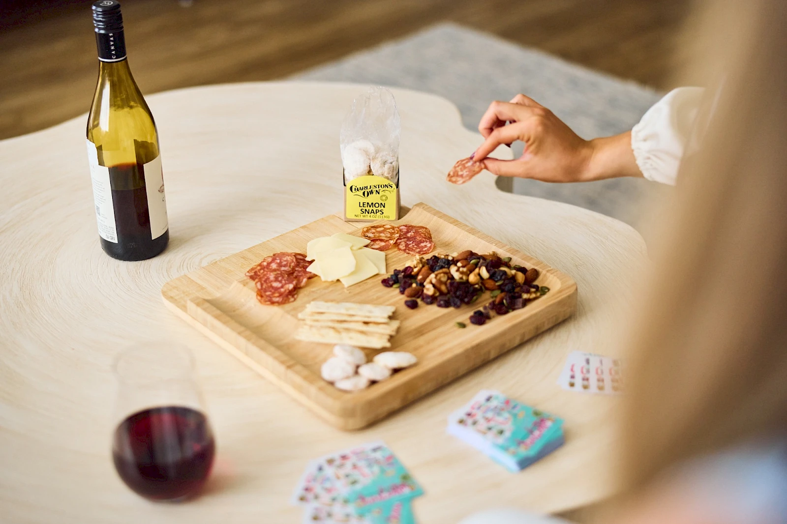 A cheese and meat board with crackers, nuts, a bottle of wine, a glass, and some cards on a table.