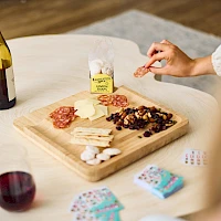 A cheese and meat board with crackers, nuts, a bottle of wine, a glass, and some cards on a table.