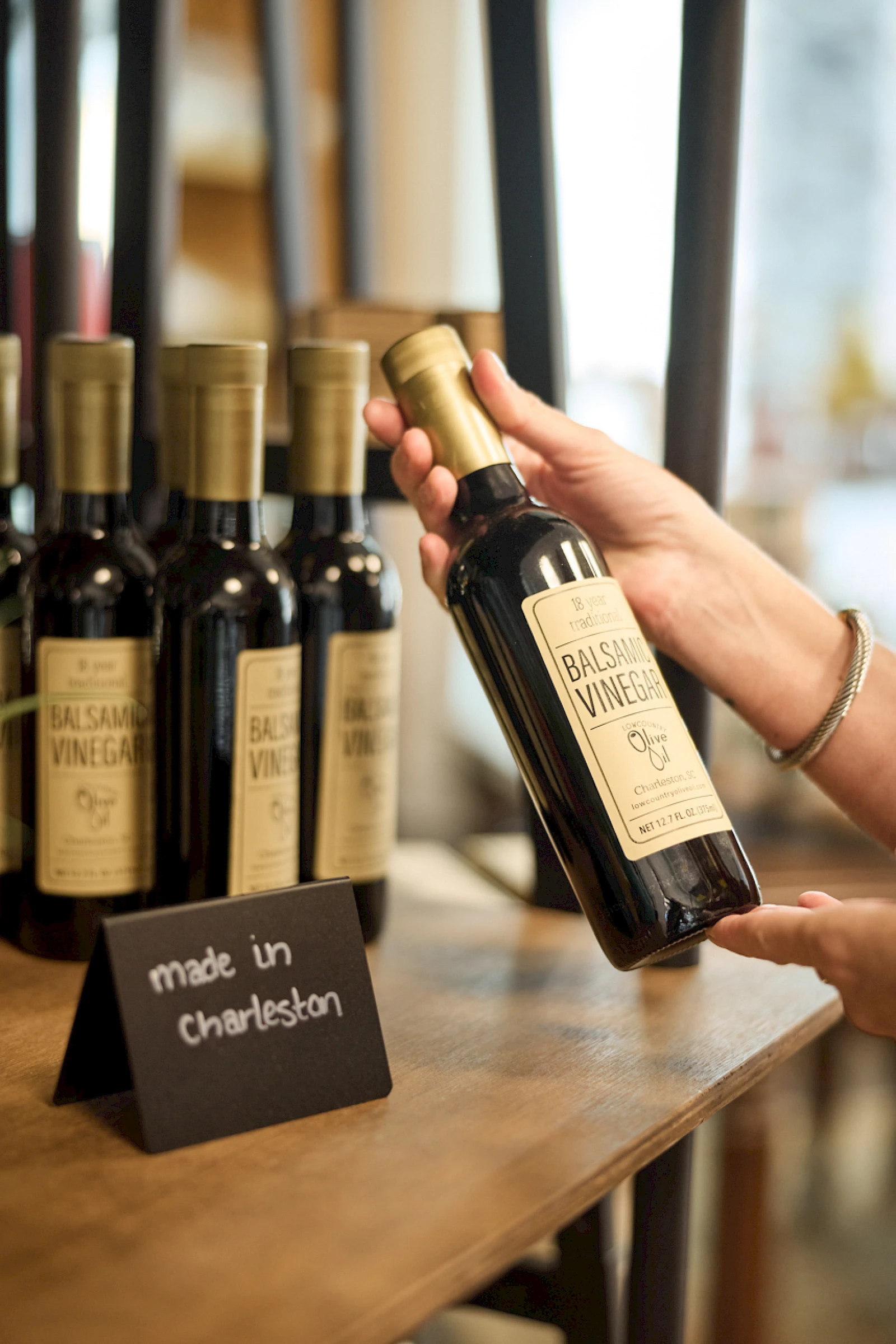 A hand selects a bottle of Brackish Vineyards wine from a row of bottles on a wooden counter, with a “made in Charleston” sign nearby.
