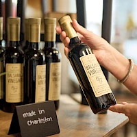 A hand selects a bottle of Brackish Vineyards wine from a row of bottles on a wooden counter, with a “made in Charleston” sign nearby.