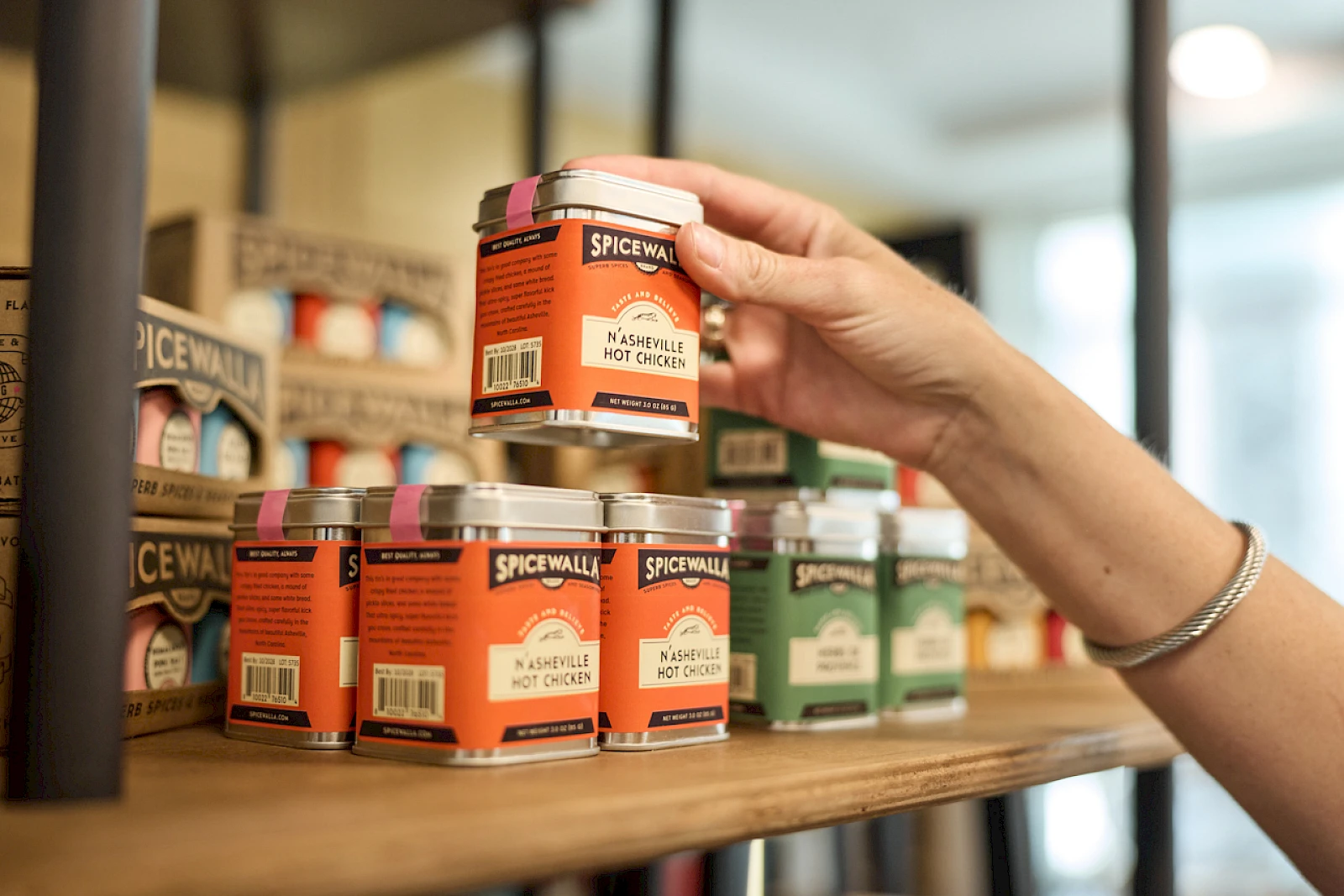 A hand selects a small orange jar labeled "Spicewand" from a shelf of colorful spice jars in a shop.
