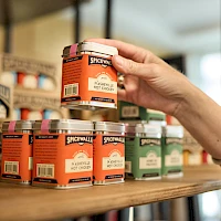 A hand selects a small orange jar labeled "Spicewand" from a shelf of colorful spice jars in a shop.