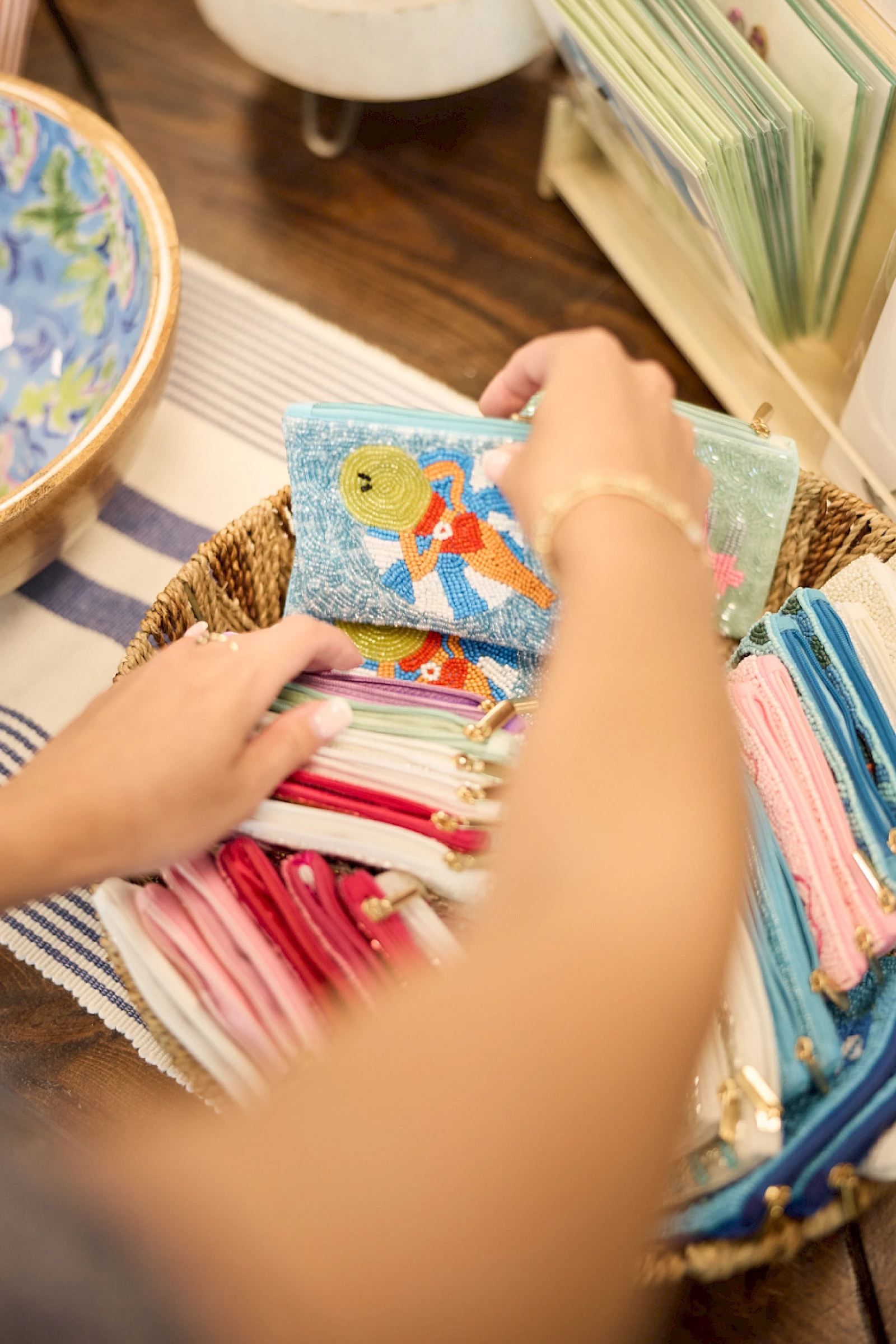 People sorting colorful fabric wallets or pouches in a basket, with a beach map and stacks of cloth nearby, inviting craft vibes indeed.