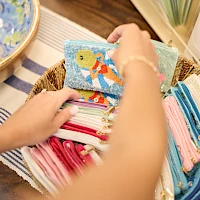 People sorting colorful fabric wallets or pouches in a basket, with a beach map and stacks of cloth nearby, inviting craft vibes indeed.