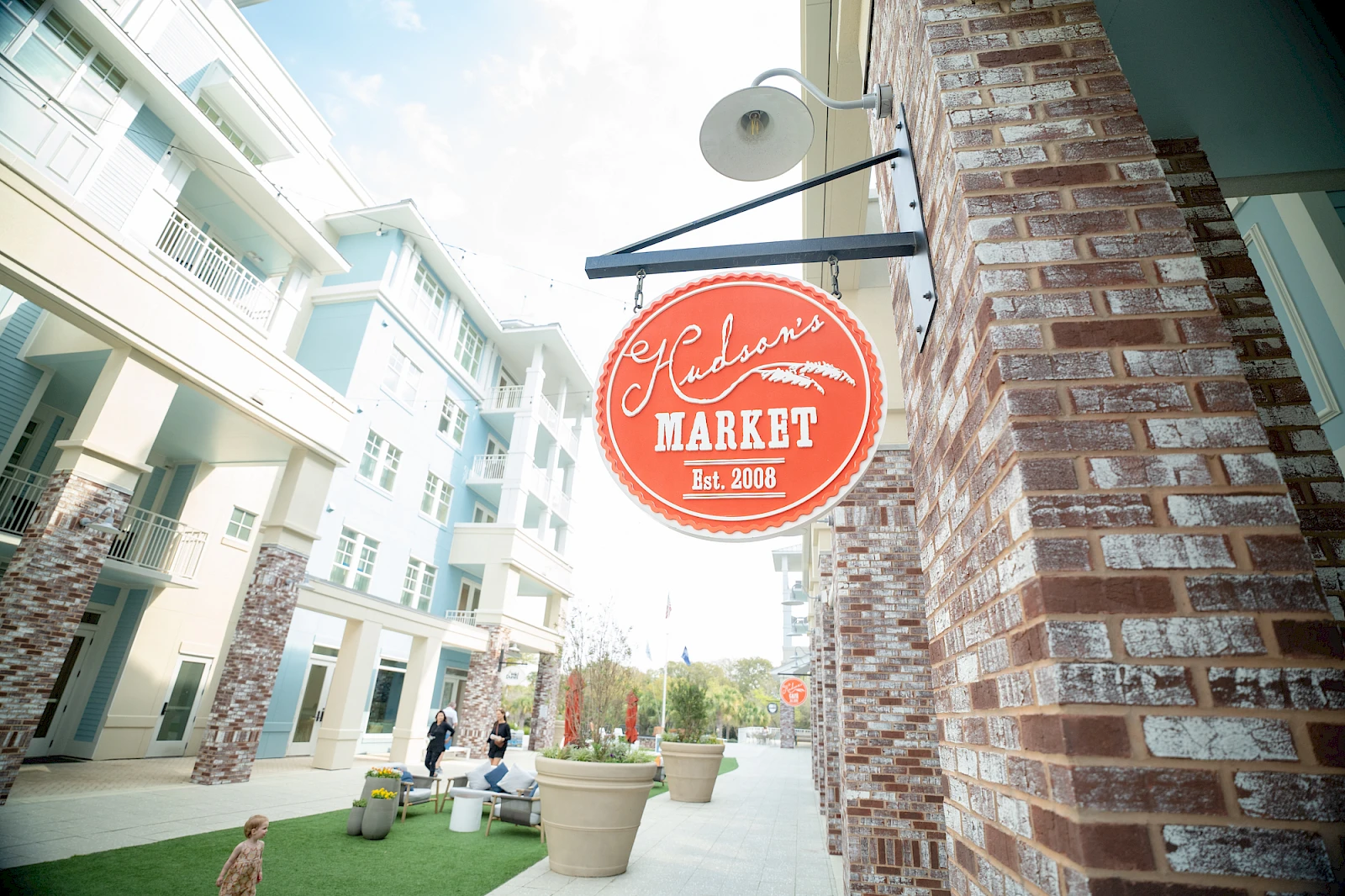 A brick storefront with a red circular sign reading “Market Est. 2006” hangs above a sidewalk, modern apartments line the street.