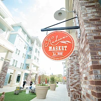 A brick storefront with a red circular sign reading “Market Est. 2006” hangs above a sidewalk, modern apartments line the street.