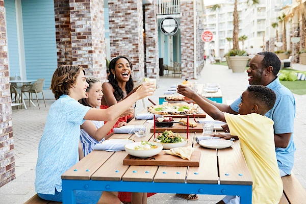 A family enjoys a meal together at a balcony table, cheering and toasting with drinks on a sunny outdoor patio.
