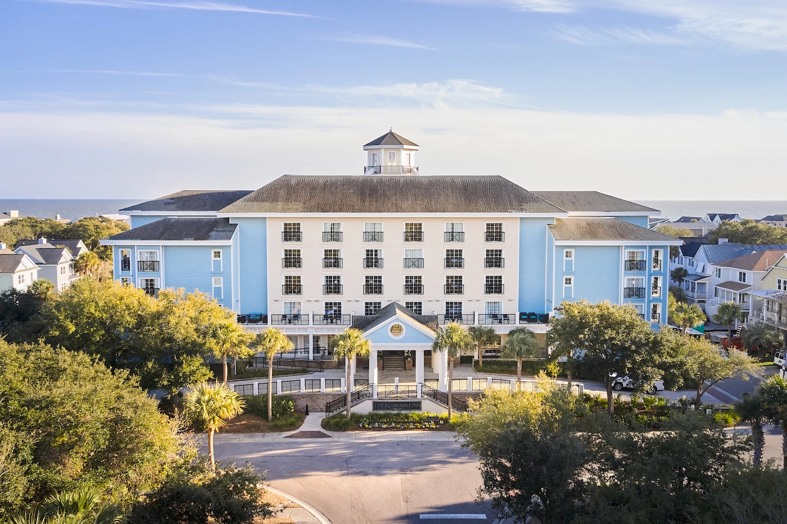 A large light-blue hotel or resort building with many windows, a central entrance, palm trees, and a scenic coastal setting in the background.