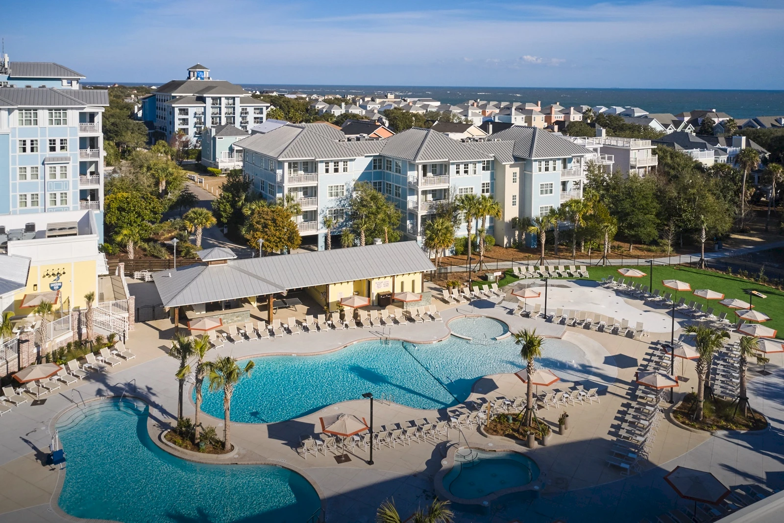 A resort pool area with lounging chairs, palm trees, and a curved blue pool surrounded by white buildings and a sunny seaside backdrop.