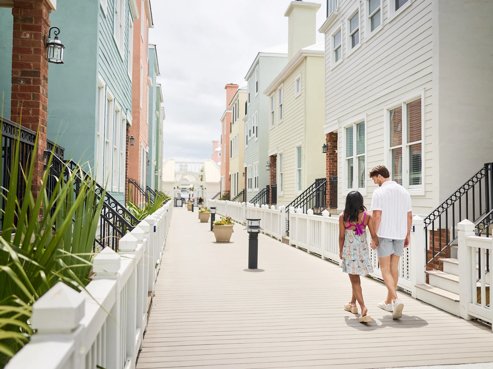 Colorful beach houses line a wooden boardwalk, a couple strolls hand-in-hand along the path, sunny skies glow over the quaint neighborhood.