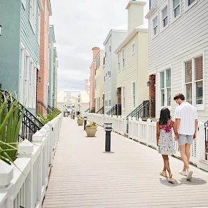 Colorful beach houses line a wooden boardwalk, a couple strolls hand-in-hand along the path, sunny skies glow over the quaint neighborhood.