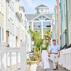 Two people walk hand-in-hand down a sunny, colorful alley lined with pastel houses and white fences. The scene feels like a charming coastal neighborhood.