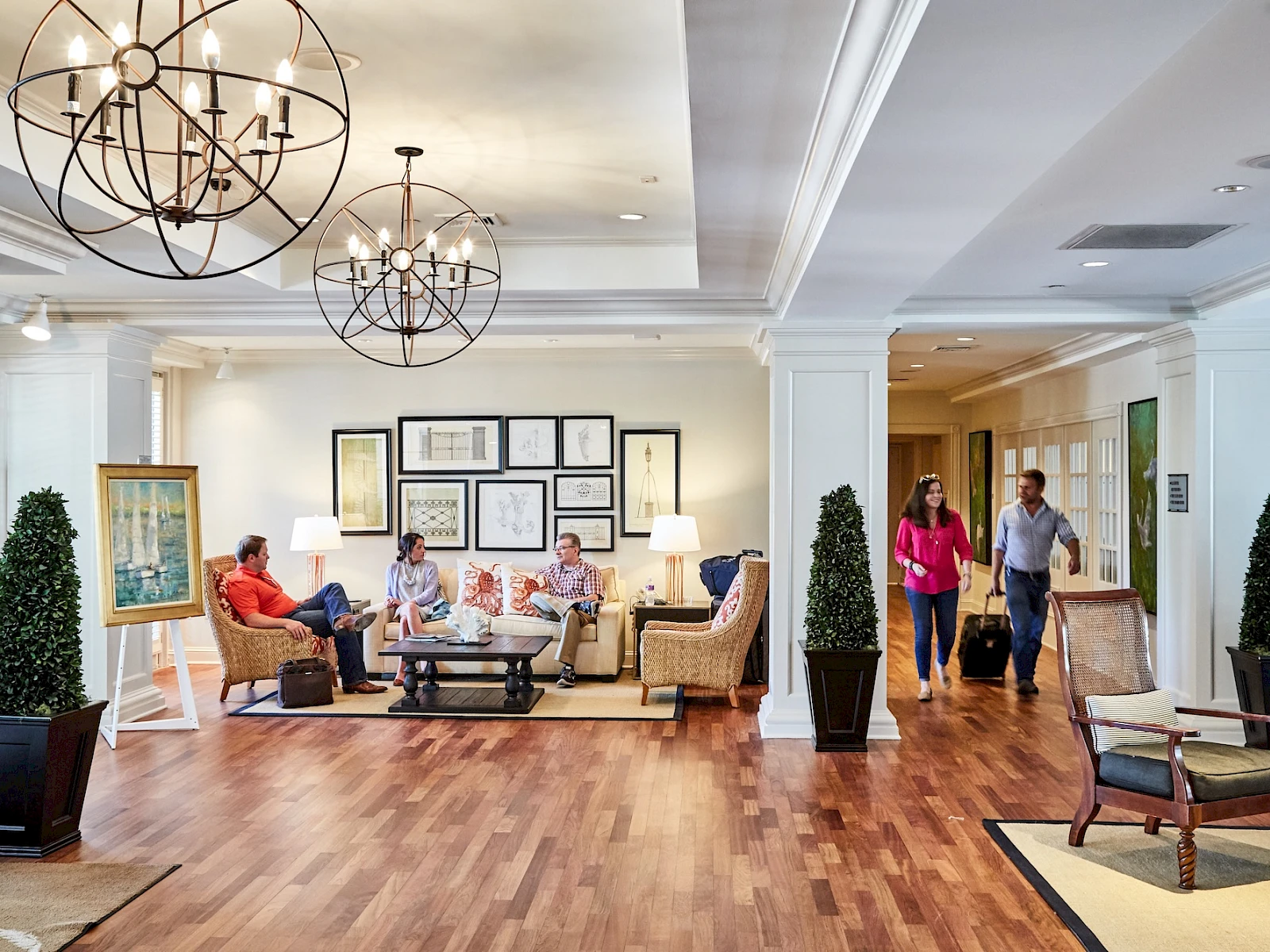 A welcoming hotel lobby with a seating area, chandeliers, wooden floors, potted plants, and guests chatting while staff assist at the reception.