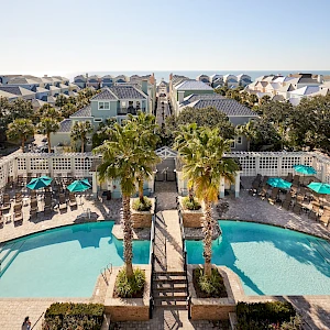 A sunny coastal pool area with clear blue water, palm trees, lounge chairs, and pastel houses in the background—resort vibes abound.