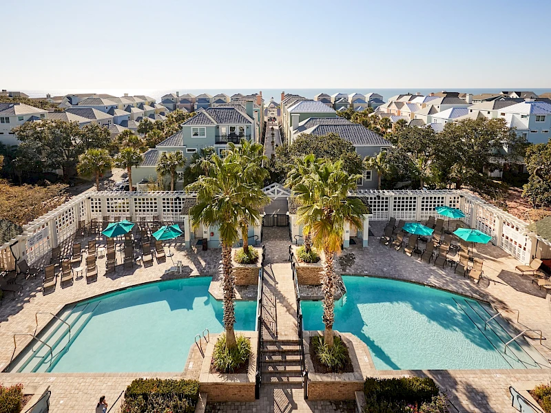 A sunny coastal pool area with clear blue water, palm trees, lounge chairs, and pastel houses in the background—resort vibes abound.