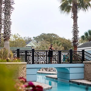 A resort pool with turquoise water, a decorative bridge, palm trees, blooming flowers, and a person standing on the bridge overlooking the pool.