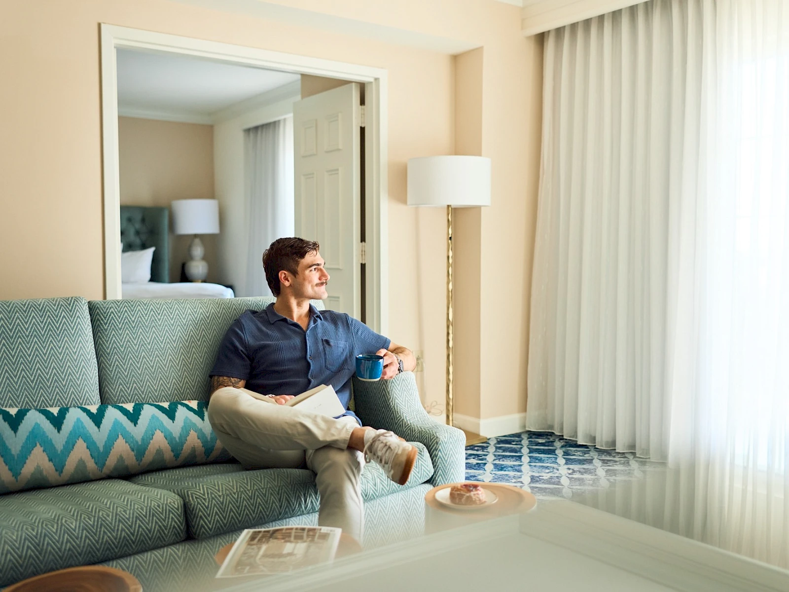 A man sits on a teal sofa in a bright hotel room, looking toward the window with sheer curtains, a lamp, and a mirror behind him.