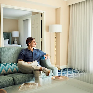 A man sits on a teal sofa in a bright hotel room, looking toward the window with sheer curtains, a lamp, and a mirror behind him.