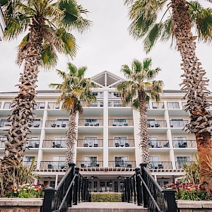 A beachfront hotel with blue and white facade, palm trees lining the entrance, and a stairway leading up to the lobby.