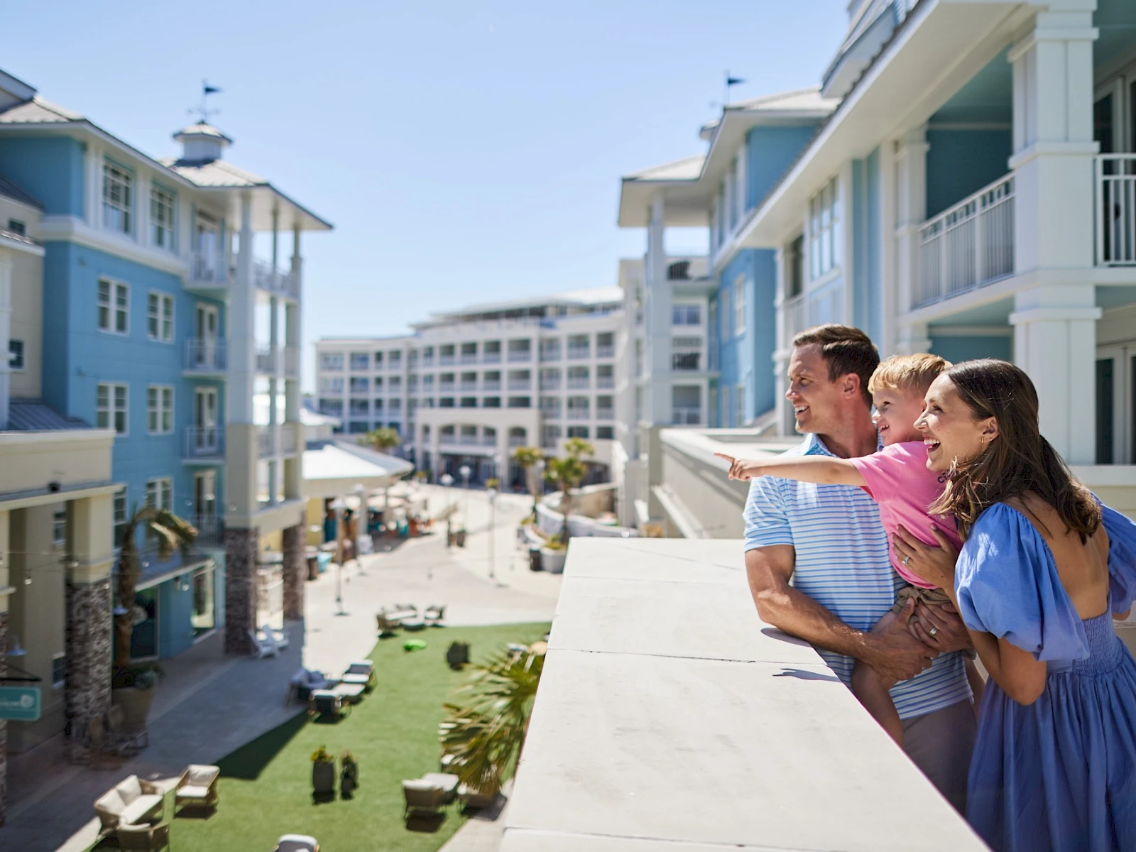 A couple on a balcony overlook a sunny coastal hotel complex with blue buildings and people strolling below.