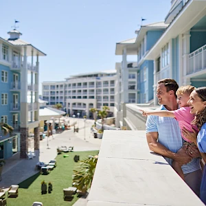 A couple on a balcony overlook a sunny coastal hotel complex with blue buildings and people strolling below.