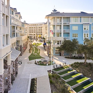 A resort-style courtyard with multi-story pastel buildings, balconies, paved walkways, green lawns, and steps leading to seating areas and trees.