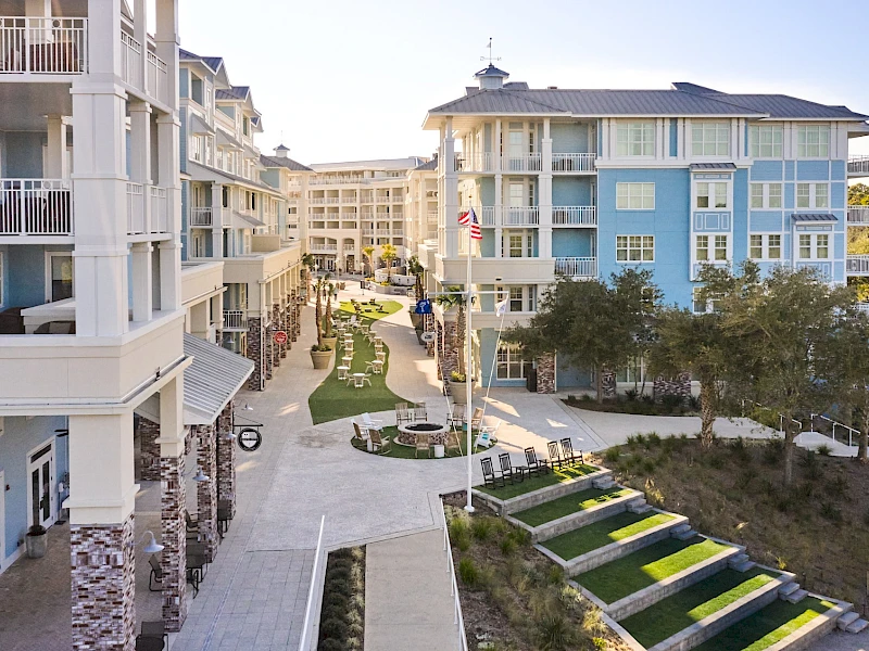 A resort-style courtyard with multi-story pastel buildings, balconies, paved walkways, green lawns, and steps leading to seating areas and trees.