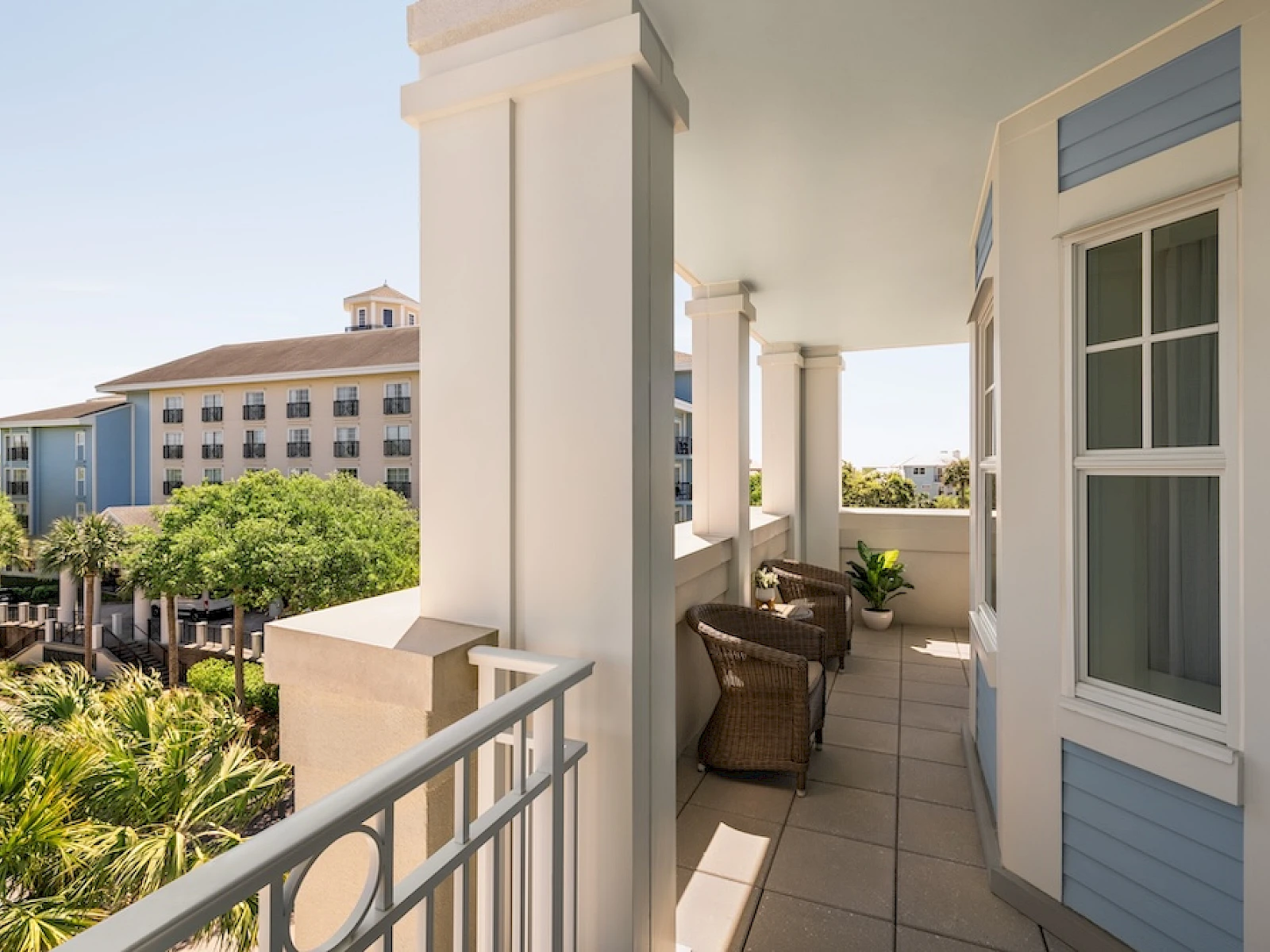 A bright seaside balcony with white columns, wicker chairs, and blue-trimmed walls overlooking palm trees and a distant hotel, sunny and inviting.