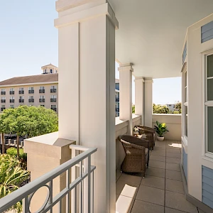 A bright seaside balcony with white columns, wicker chairs, and blue-trimmed walls overlooking palm trees and a distant hotel, sunny and inviting.