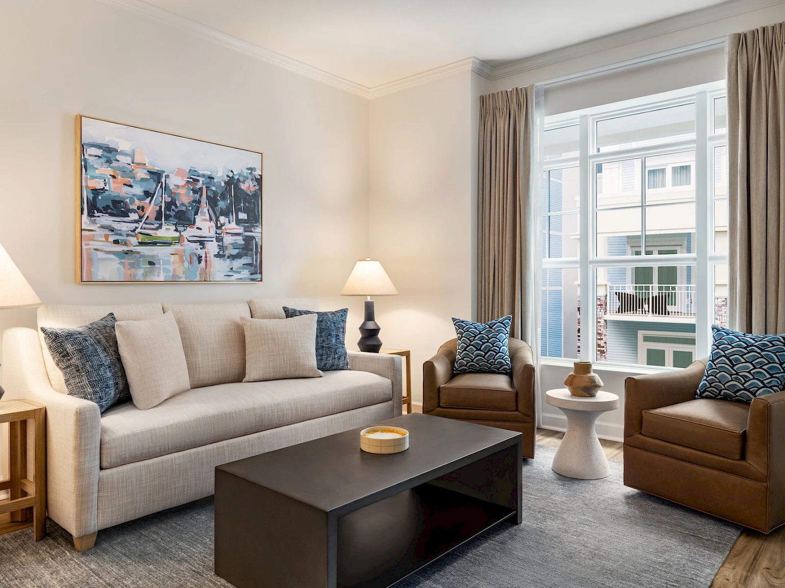 A bright, modern living room with a beige sofa, two brown armchairs, a dark coffee table, art on the wall, and large windows with curtains.
