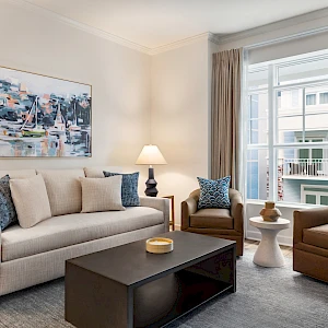 A bright, modern living room with a beige sofa, two brown armchairs, a dark coffee table, art on the wall, and large windows with curtains.