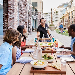 A family dinner outdoors on a long table, serving pizza and sides on a sunny street with kids and adults chatting warmly, at a resort courtyard.